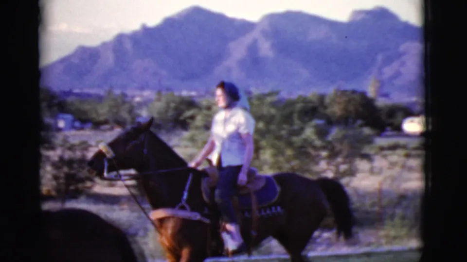 A woman rides a dark horse through a rural Cottonwood, Arizona setting, with scrub, distant mountains, and a pale vehicle visible behind her.