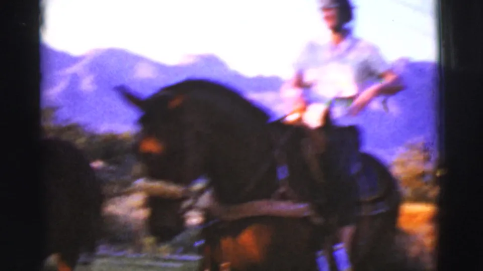 Soft 8mm frame of a woman riding a dark horse outdoors, with Arizona mountains and scrub vegetation behind her.