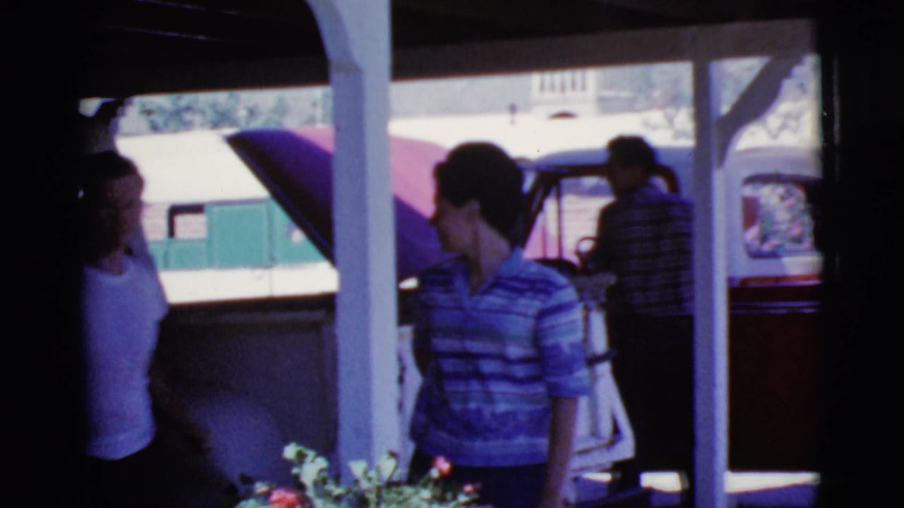 People stand under a shaded carport near parked cars and flowering shrubs.