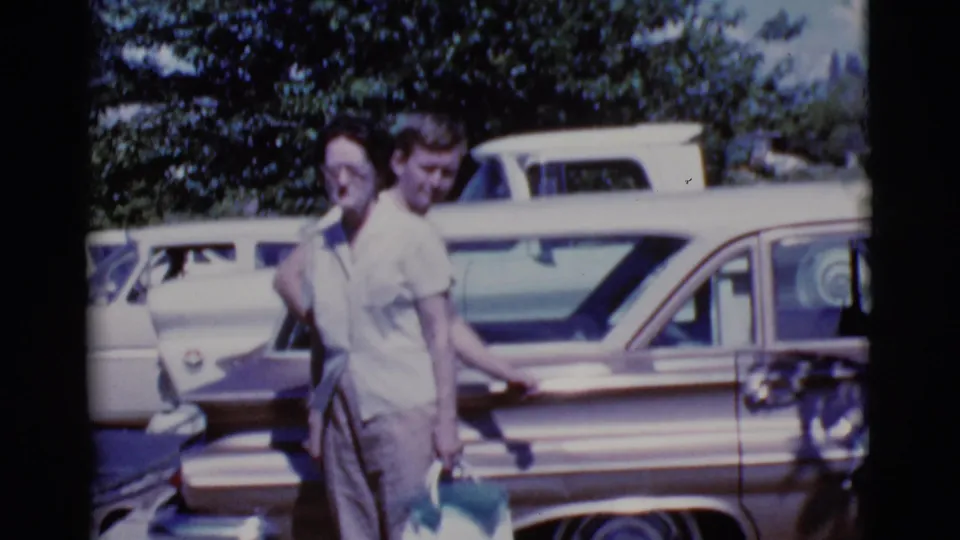 In a 1968 Cottonwood, Arizona home movie, a woman carrying a purse walks with a man beside mid-century cars.