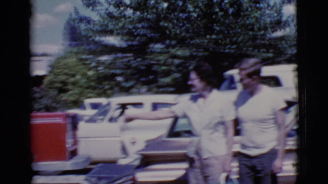 Two adults walk near parked cars in a sunny lot, with trees behind them.