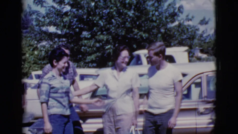 Three adults interact beside a brown car in the 1968 Cottonwood parking-lot scene, with another white vehicle behind them.