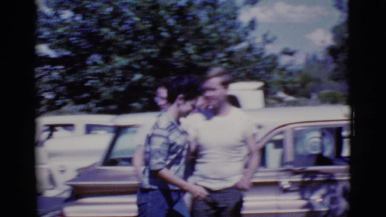Two adults stand near a parked station wagon while another person is partly visible behind them.