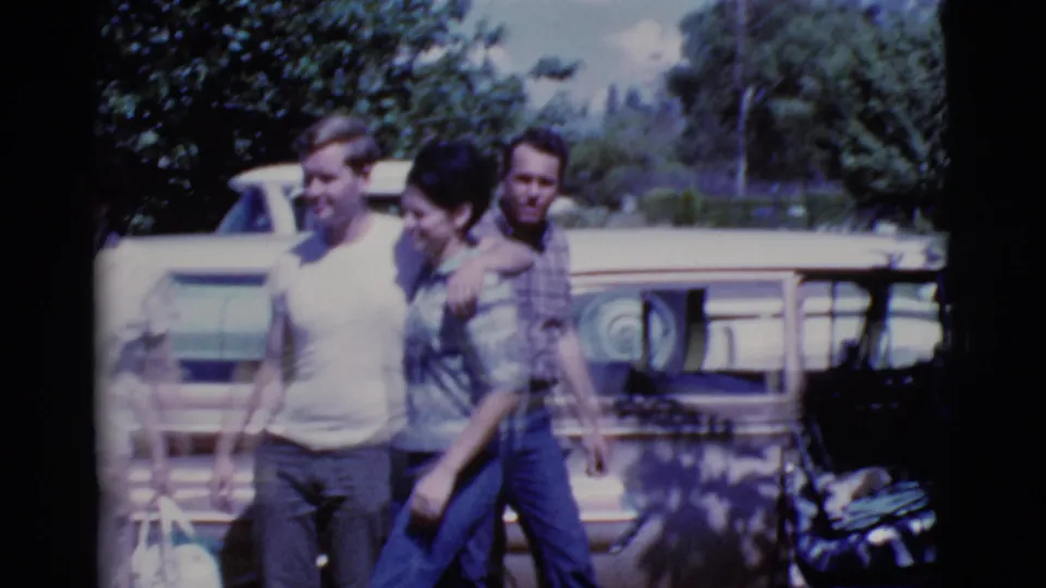 Three adults in casual 1960s clothing move together in a Cottonwood, Arizona parking lot with a station wagon behind them.