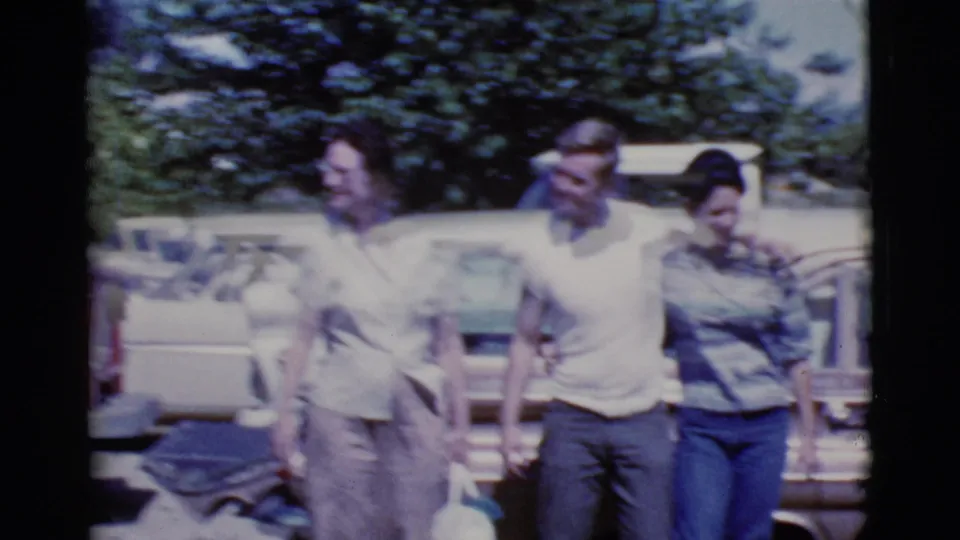 A blurred 1968 home-movie frame shows adults grouped beside cars in a sunny Cottonwood, Arizona parking lot.