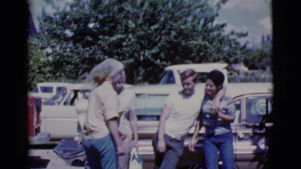 A woman faces a small group of adults beside parked cars in a sunny 1968 Cottonwood, Arizona home-movie scene.