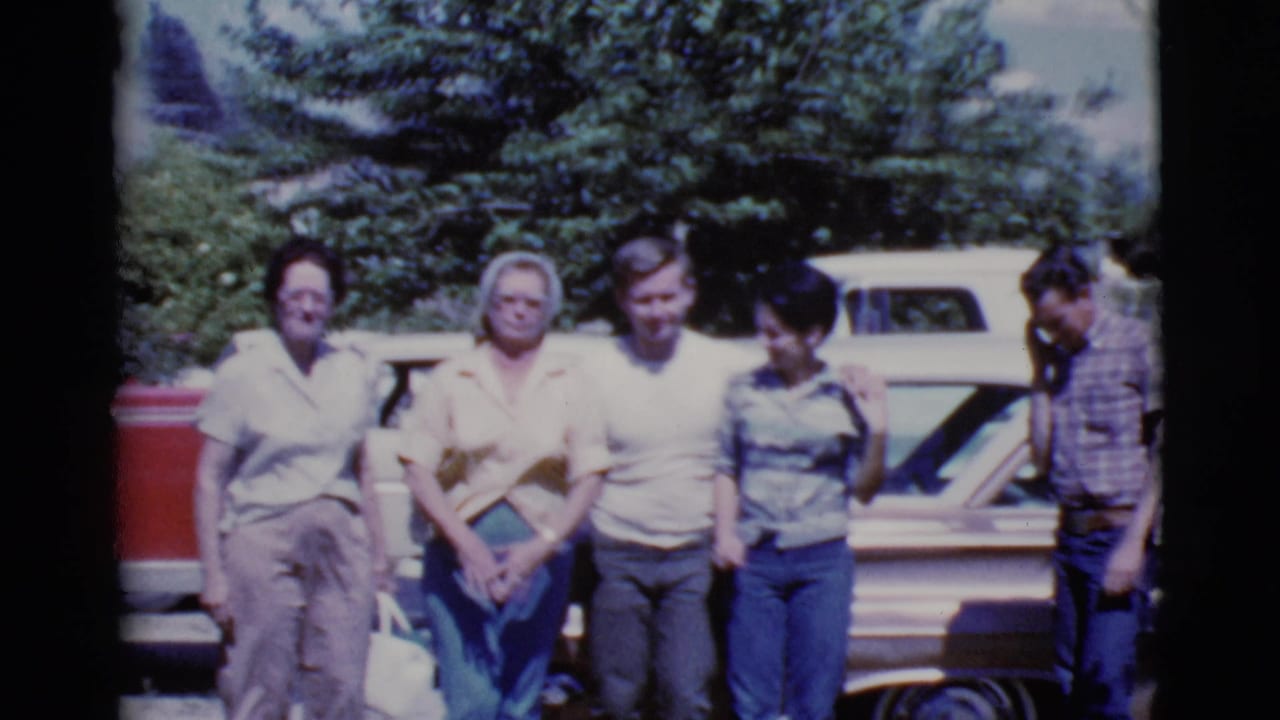 Five people stand in front of a brown car with trees and a red vehicle behind them.