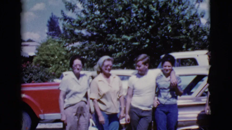 Four people in casual 1960s clothing stand by a brown car in the Cottonwood, Arizona home-movie scene, smiling and turning toward one another.
