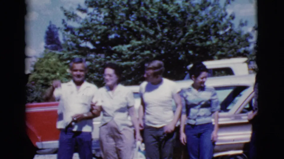 Adults in casual 1960s clothing walk together beside parked vehicles in Cottonwood, Arizona, with trees filling the background.