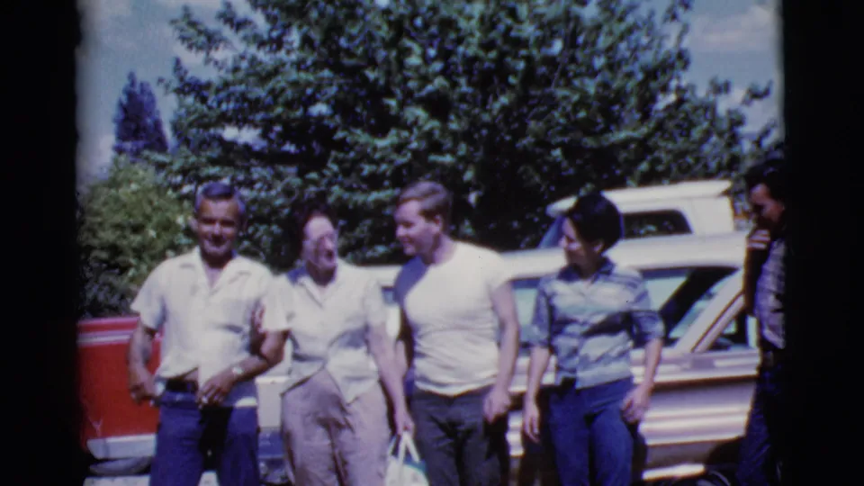 The group continues walking and talking beside a red vehicle and pale car, with 8mm softness and saturated color visible.