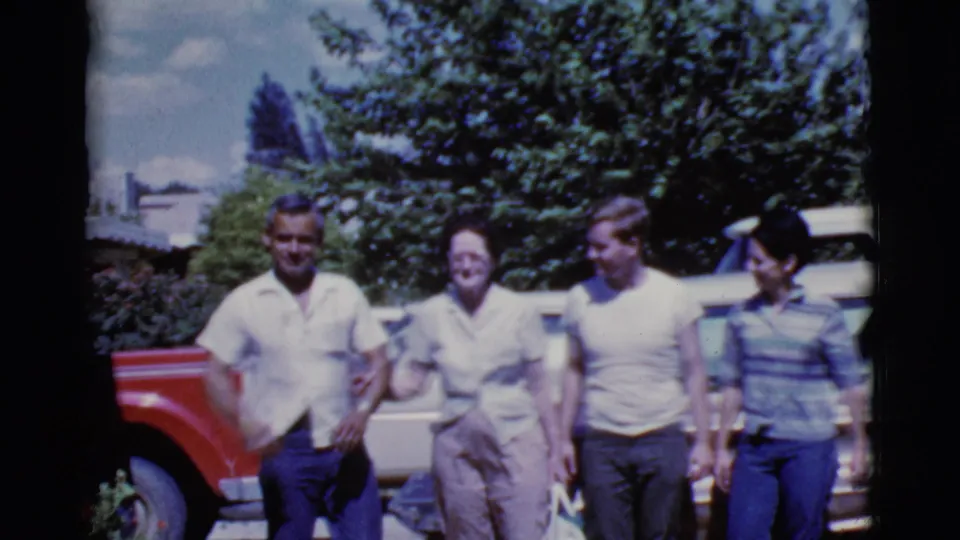 Adults walk closely together near a red vehicle and light-colored car in the metadata-supported 1968 Cottonwood home-movie scene.