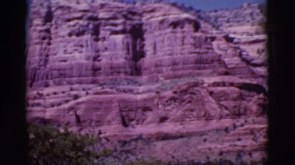 A closer 8mm frame of the Arizona red rock face shows vertical staining, horizontal strata, and low shrubs at the canyon base.