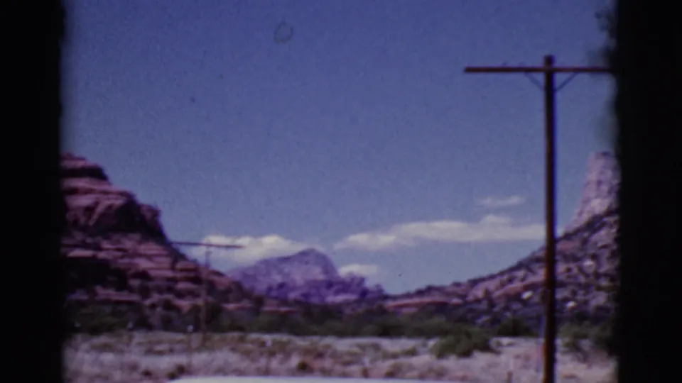 1968 Arizona desert view with a foreground utility pole, red rock canyon edges, low scrub, distant mountain, and blue sky.