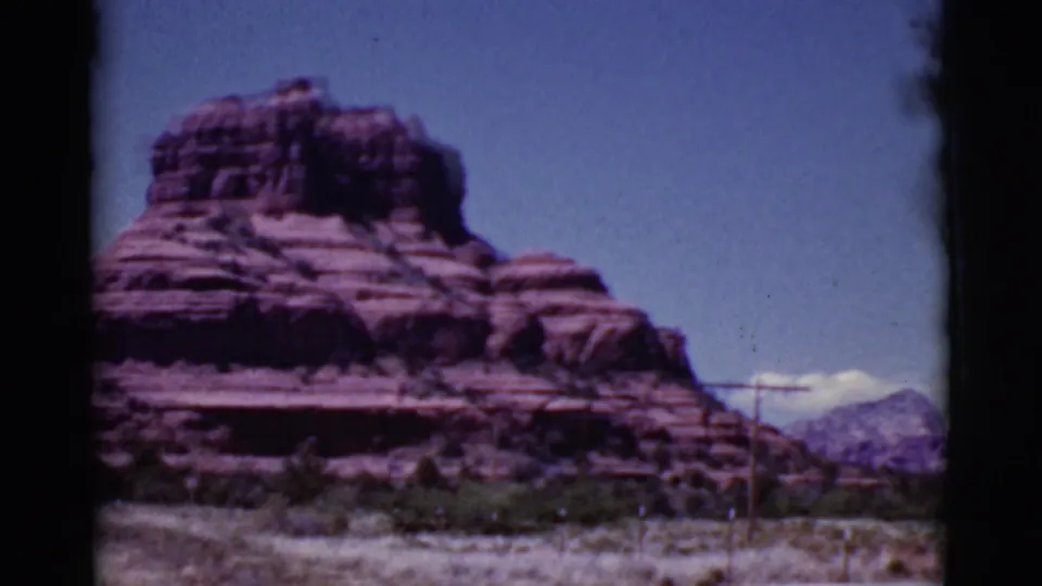Cottonwood-area 8mm frame showing a broad red rock butte, sparse desert vegetation, utility poles, distant hills, and blue sky.