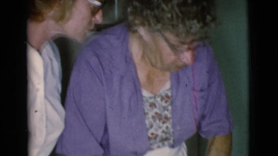 A woman in a white shirt leans close beside an older woman in purple during a quiet indoor 1968 Cottonwood home-movie moment.