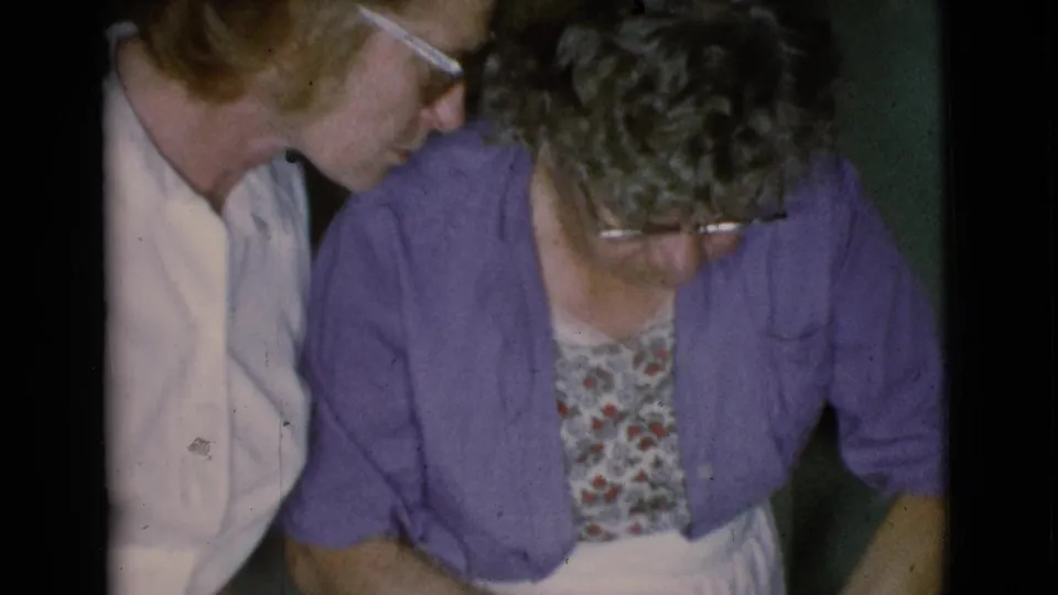 The older woman in purple bends forward as the second woman leans near her shoulder in this brief 1968 domestic home-movie frame.