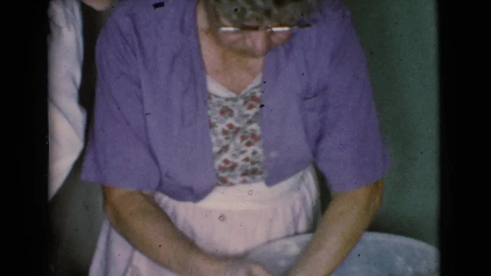 In 1968 Cottonwood, Arizona footage, an older woman in a purple cardigan and apron leans over a bowl while handling clay.