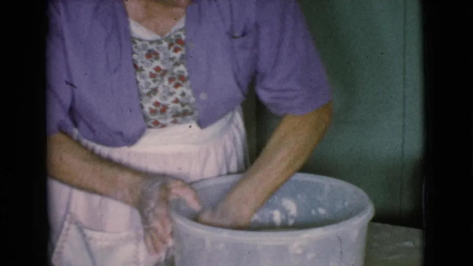 The woman stands beside a large bowl, pressing a clay-covered hand into the material in a home interior.
