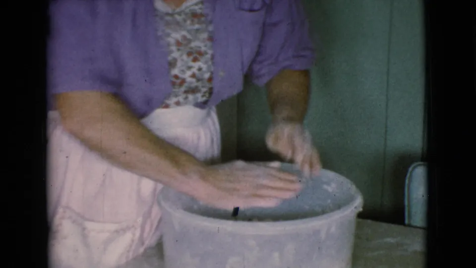 An older woman in a purple cardigan and apron holds clay-coated hands over a large bowl in the 1968 home-movie scene.