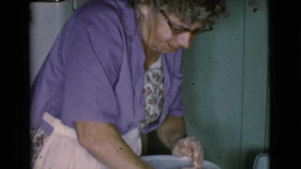 The woman leans over the work surface, focused on shaping clay beside a large bowl in a private domestic setting.