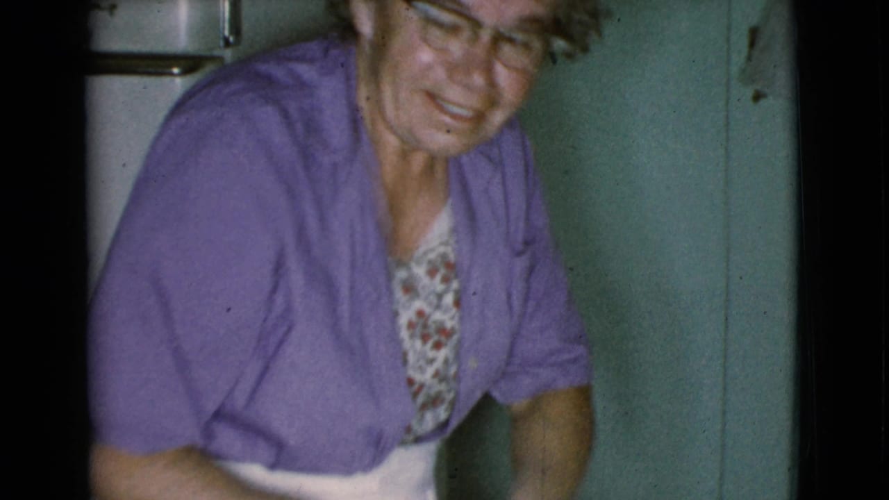 Older woman with glasses in a purple cardigan sits or leans in a kitchen.