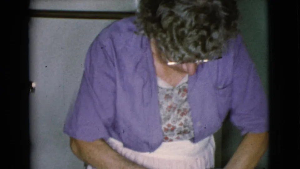 The woman bends forward over a work area, wearing a purple cardigan and patterned dress in the Cottonwood kitchen interior.