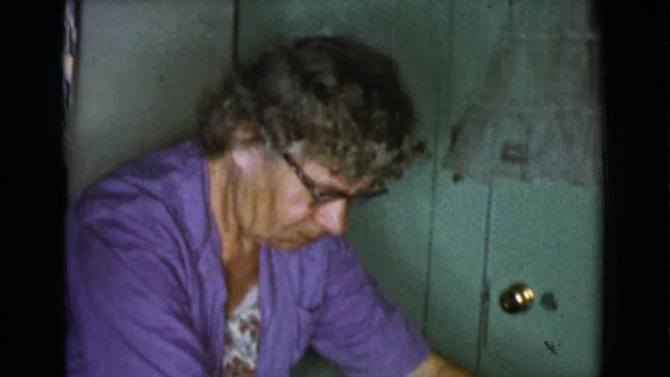 In profile, the woman continues working at close range beside a green kitchen wall or cabinet, with film grain and dark frame edges visible.