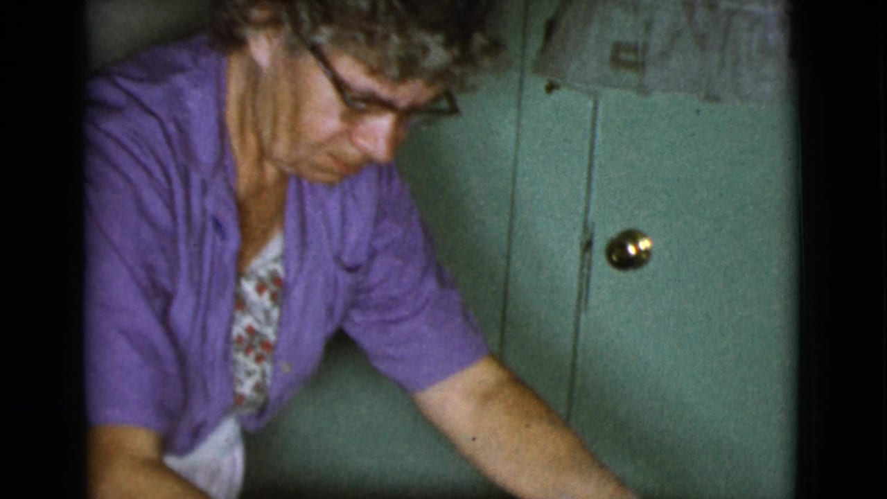 Woman in a purple cardigan leans over in a kitchen near a green cabinet door.