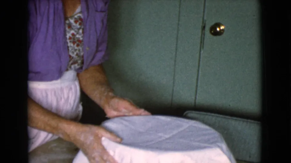 A woman in a purple cardigan smooths a white cloth over a bowl, with flour visible on her hands in a private kitchen setting.