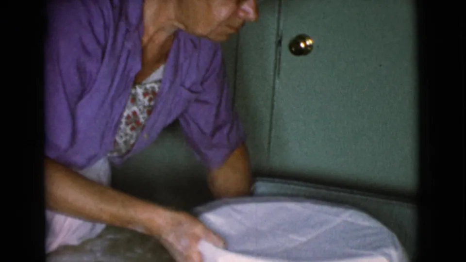 The woman continues adjusting the cloth over the bowl, her purple cardigan and floral blouse visible against the green cabinet background.