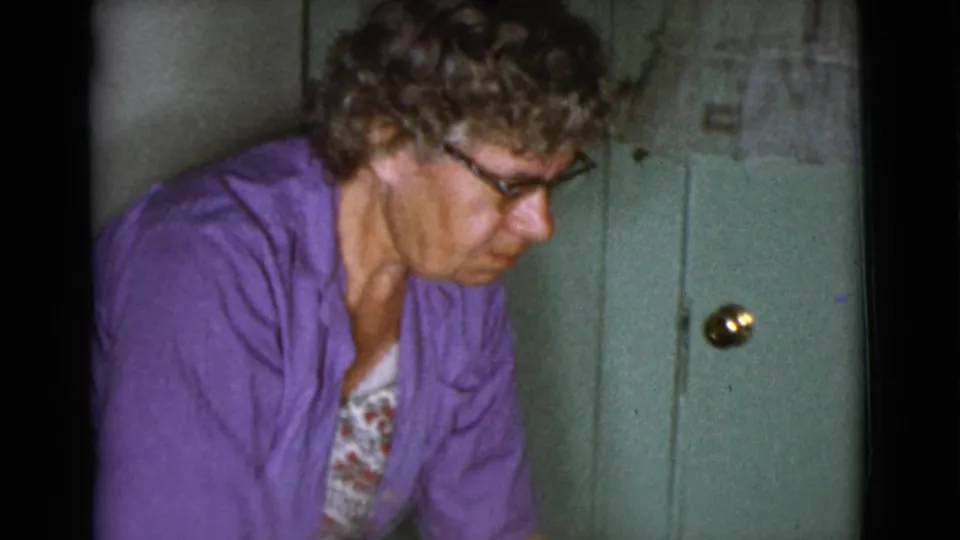 A woman in glasses and a purple cardigan leans forward in the kitchen, focused downward as the home-movie camera frames her close to a green cabinet.