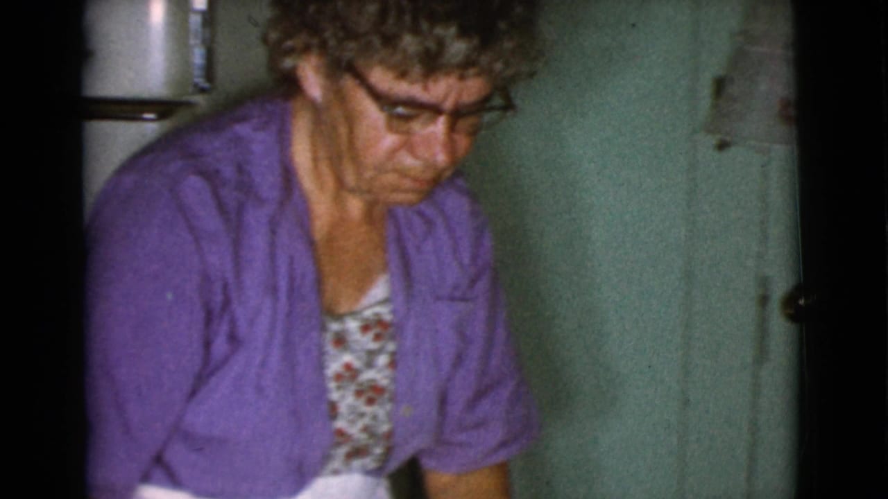 Woman in glasses and purple cardigan seated or leaning in a kitchen.