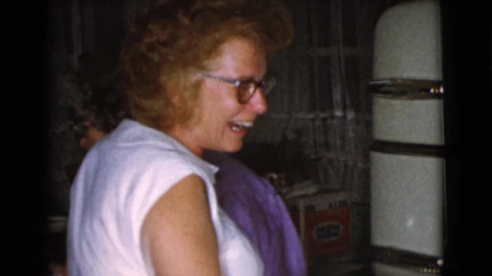 A woman in a white sleeveless top smiles in the kitchen, with another woman partly visible behind her near the refrigerator.