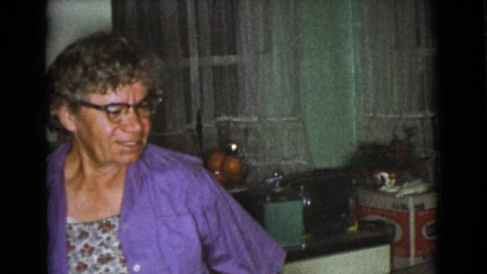 The woman in glasses and a purple cardigan looks across the 1968 Cottonwood kitchen, with a sink, curtains, and countertop items behind her.