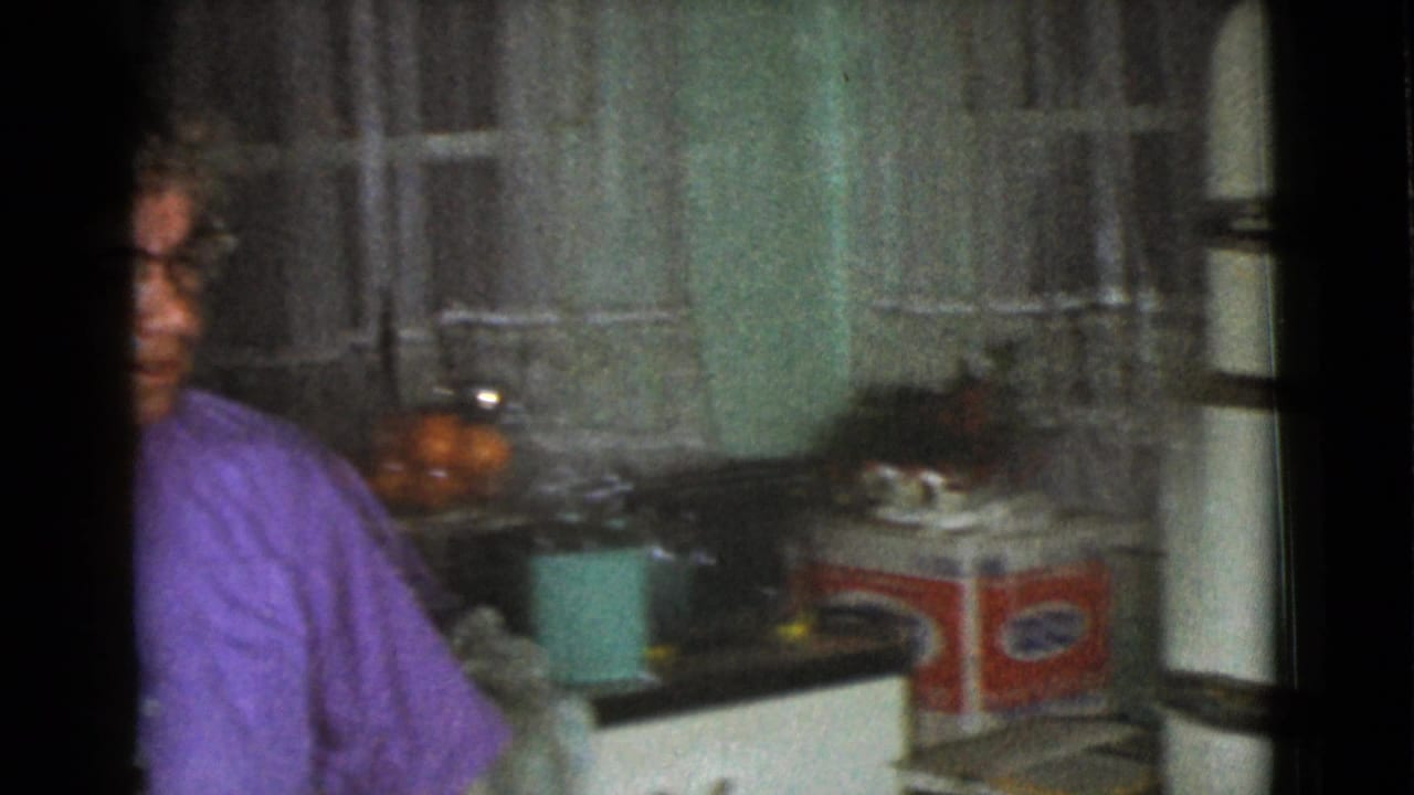 Older woman in a purple top sits beside a kitchen counter and sheer curtains.