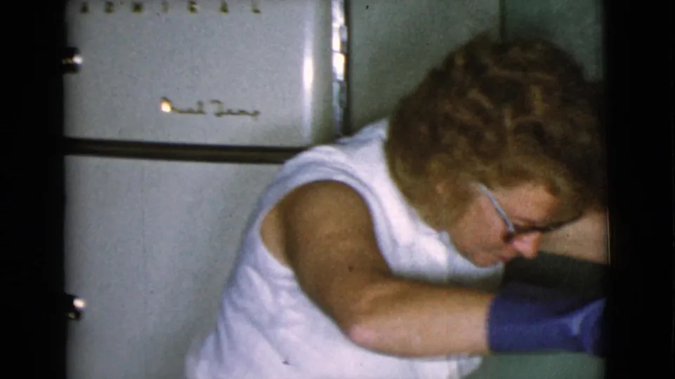 In the Cottonwood kitchen, an older woman wearing glasses, a sleeveless white top, and blue gloves leans near a white refrigerator.