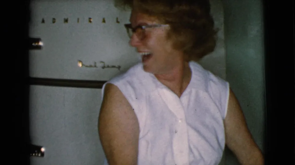 An older woman laughs and looks to the side in a 1968 Cottonwood kitchen, with a white refrigerator visible behind her.