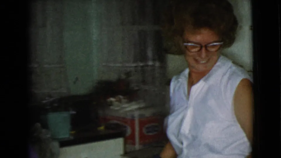 The woman smiles while standing near a kitchen counter, sheer curtains, and household items in the 1968 Cottonwood home interior.