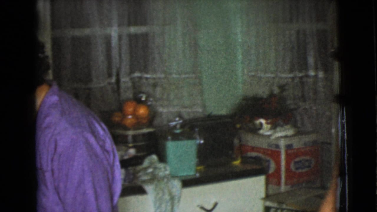 1968 Cottonwood home-movie frame showing a person in purple at the edge of a lived-in kitchen with oranges, curtains, and countertop appliances.