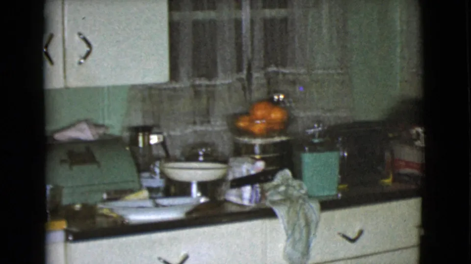 1968 Cottonwood kitchen detail with oranges, stacked dishes, green appliance, towel, pale cabinetry, and lace curtains in soft 8mm focus.