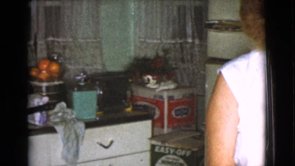 1968 Cottonwood home-movie frame with an adult in a white sleeveless blouse beside a cluttered kitchen counter and visible red product box.