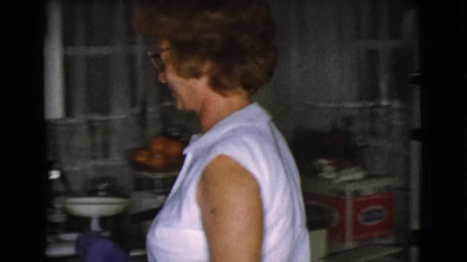 1968 Cottonwood home-movie frame showing a woman in glasses and a sleeveless white blouse in a private kitchen with oranges and dishes nearby.