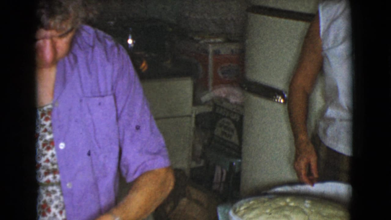 Older woman in a purple shirt stands at a kitchen work area with another adult partly visible nearby.