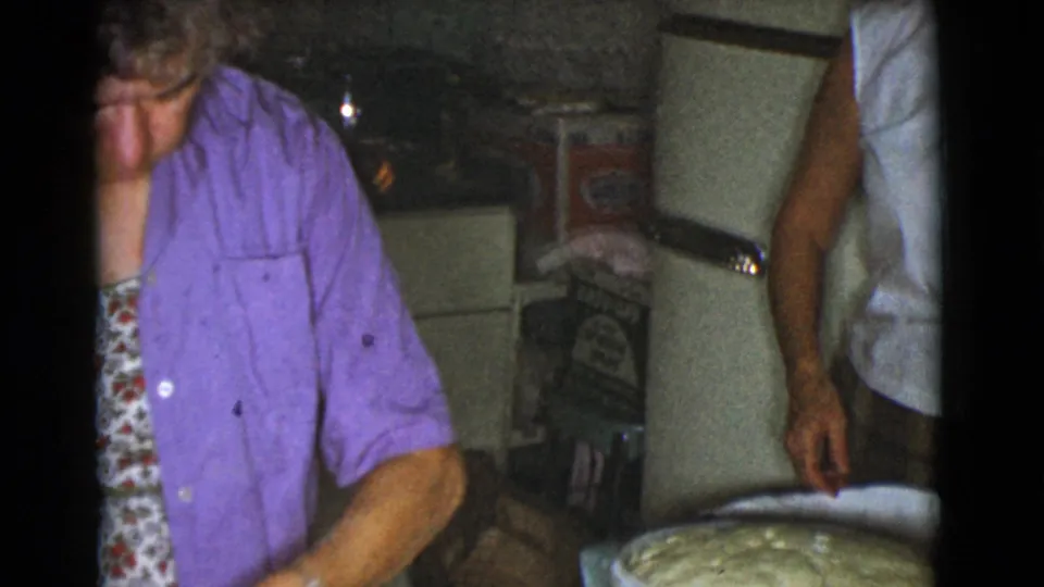 In the supplied 1968 Cottonwood, Arizona context, an older woman in a purple shirt works in a dim home kitchen as another adult stands nearby.