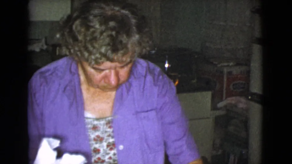 The woman looks down toward her work surface in a dim kitchen, with possible food packaging and household items behind her.