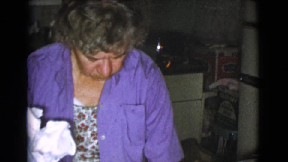 An older woman in a purple shirt leans over a kitchen surface during food preparation, with a white cloth or paper near the foreground.