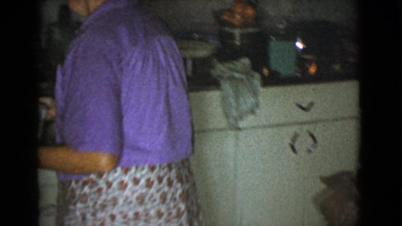 Woman in a purple top and floral apron stands in a kitchen near cabinets.