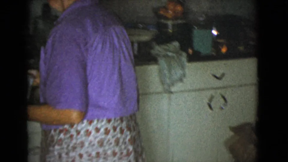 In a 1968 Cottonwood, Arizona home kitchen, a woman in a purple top and floral apron stands beside cream cabinets.