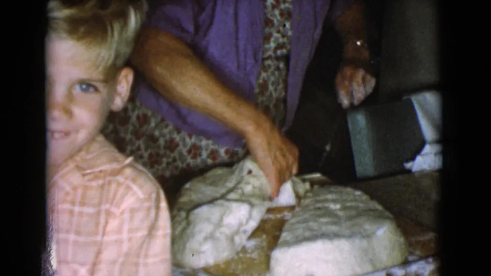 A woman divides bread dough on the wooden board as a child leans toward the camera in the 1968 home kitchen.