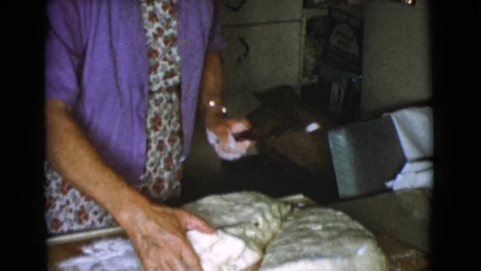 The woman continues preparing bread dough in a private kitchen, with cabinets and household items behind her.